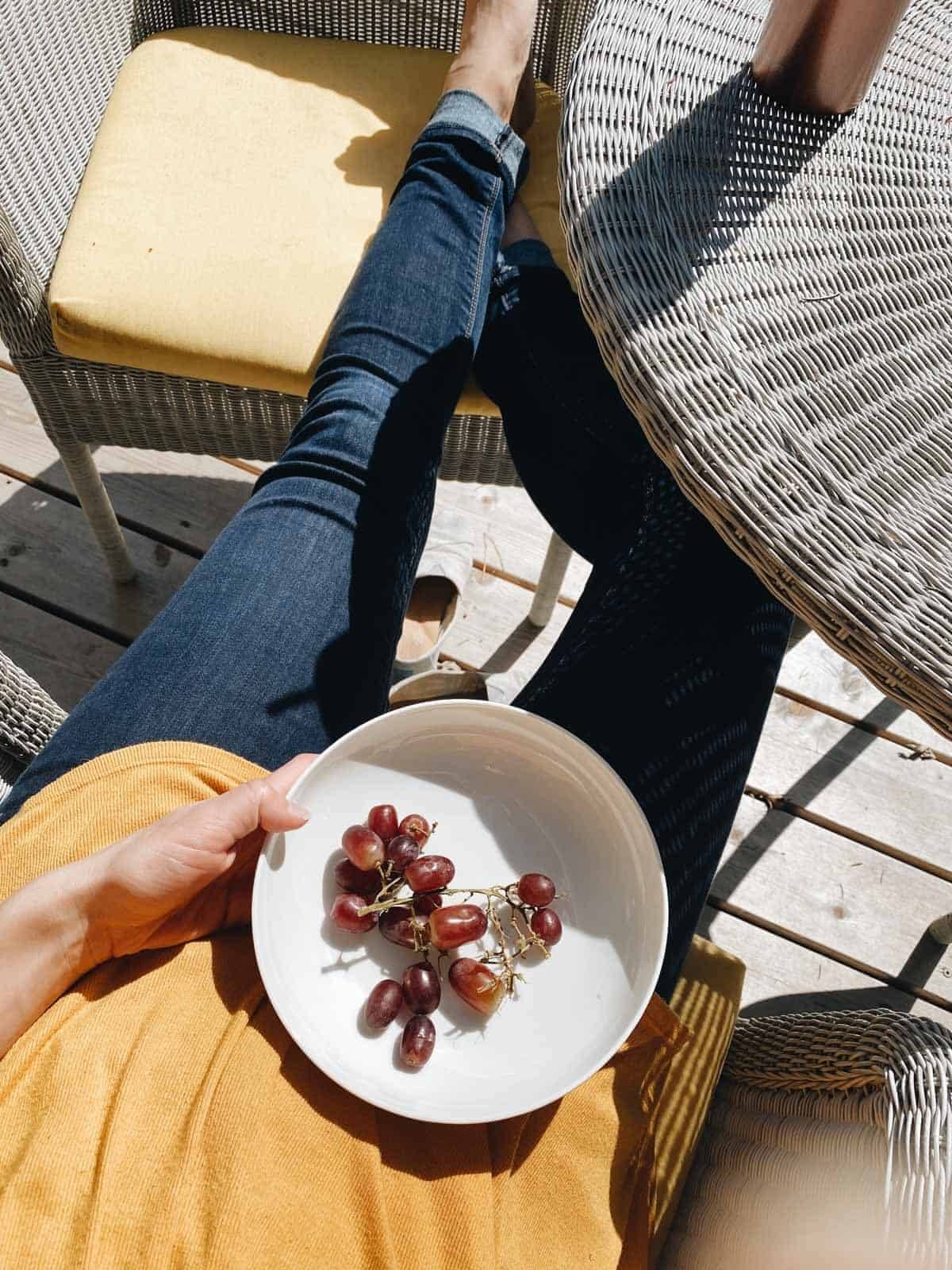 A person sitting on a chair wearing yellow top and blue jeans holding a plate having some grapes.