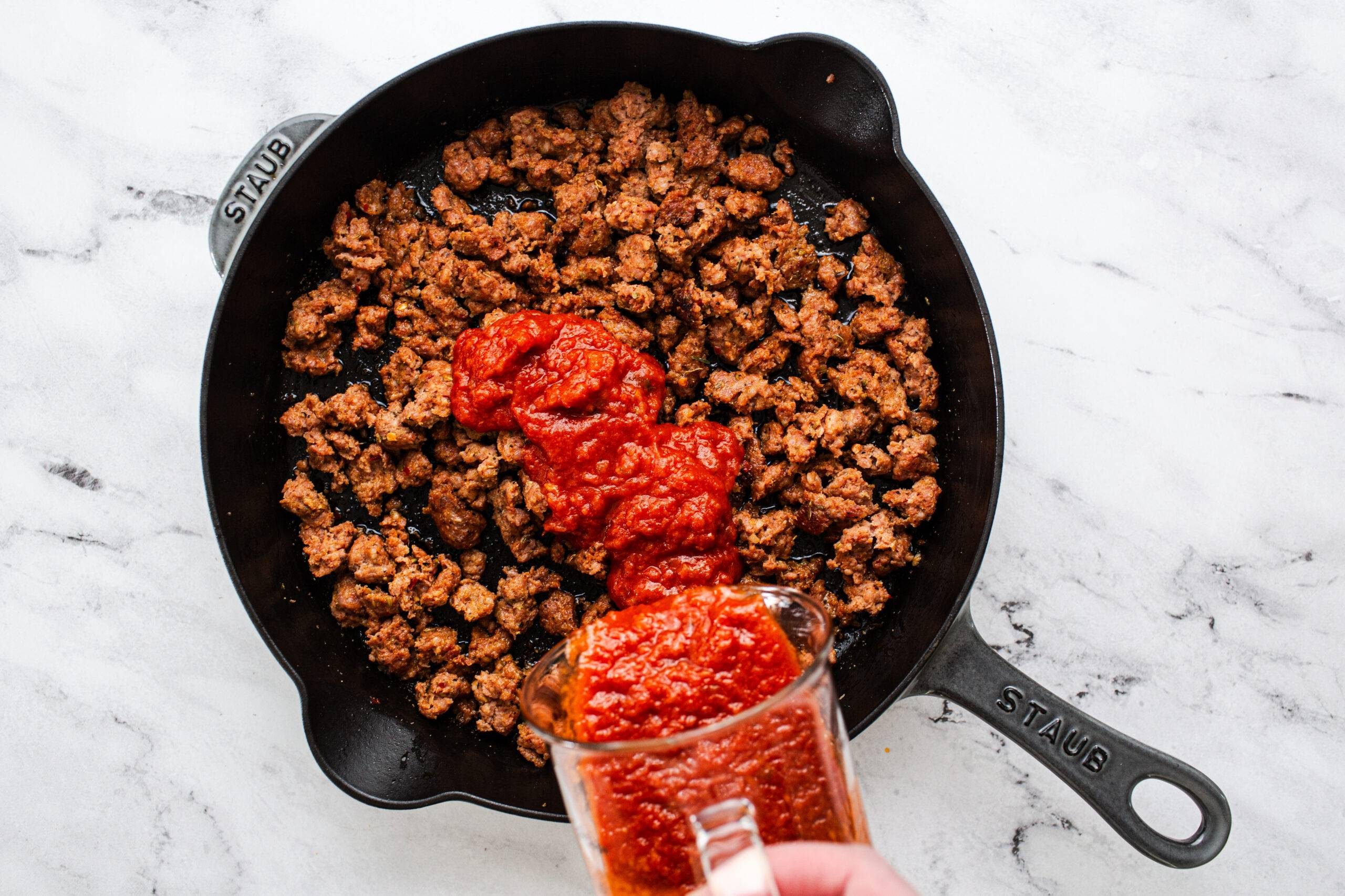 Adding marinara sauce to cooked sausage in a pan.