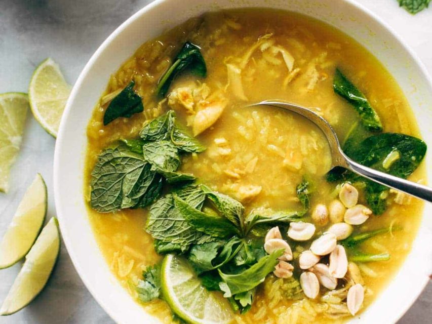 Chicken and rice soup in a bowl with a spoon. There are fresh herbs on top and lime wedges on the side.