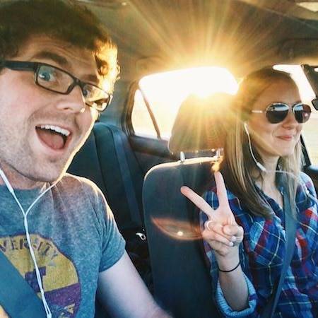 A man and a woman smiling, seated in a car, he in regular glasses, she in sunglasses, flashing the peace sign.