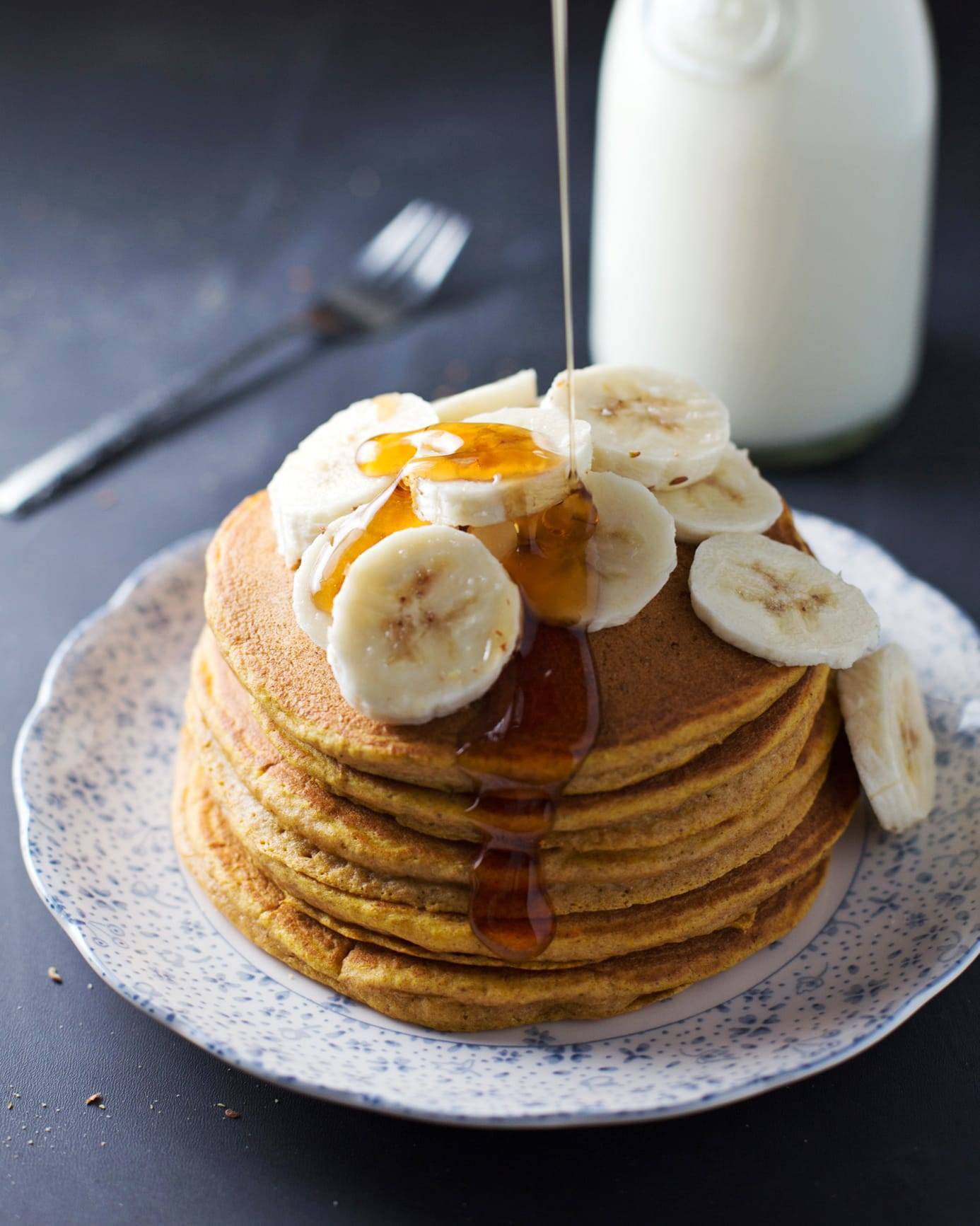 Whole wheat pumpkin pancakes stacked on a plate with bananas and syrup.