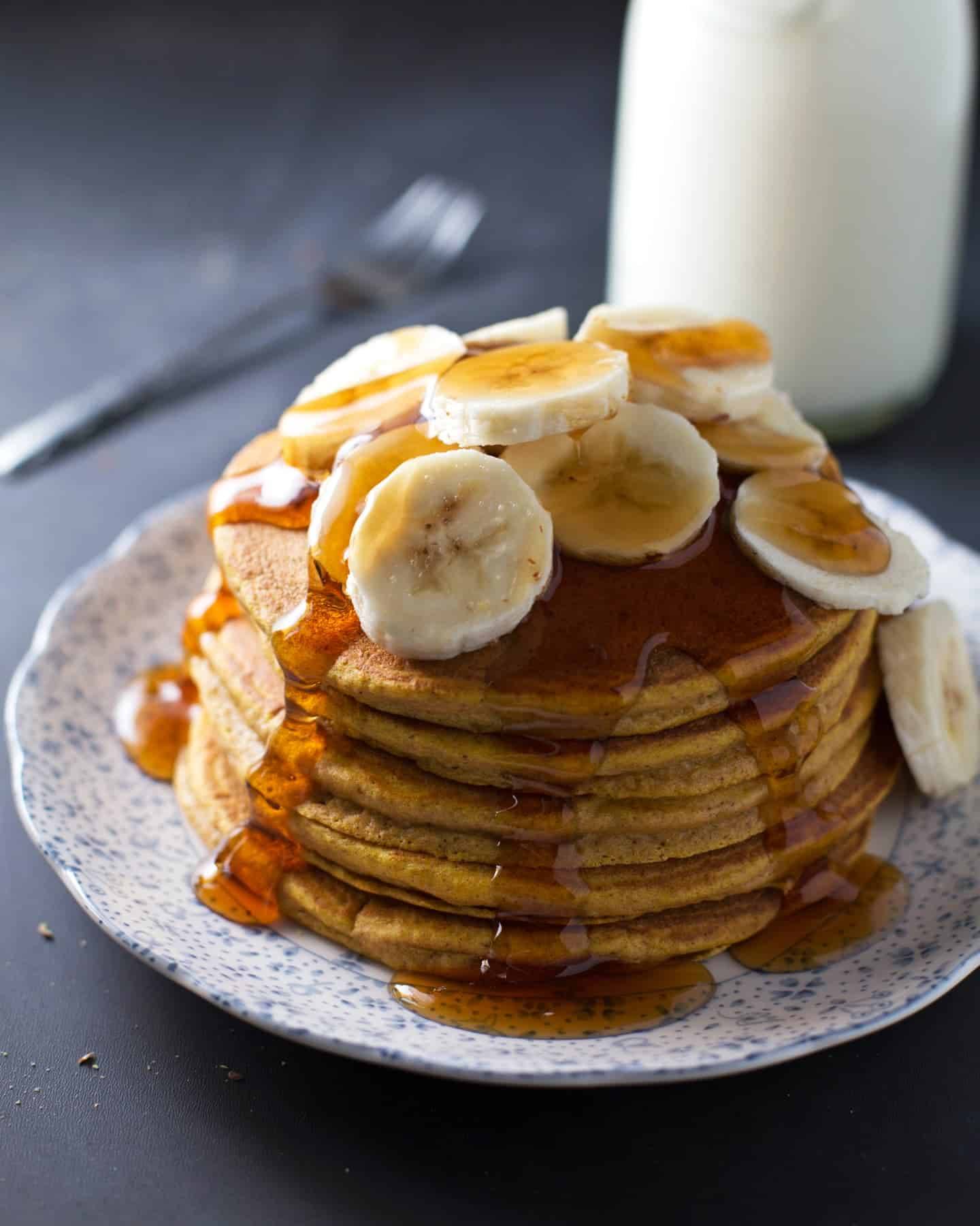 Whole wheat pumpkin pancakes stacked on a plate with bananas and syrup.