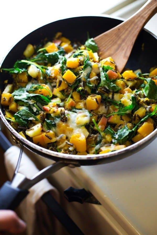 Apples, squash, sauteed onions, wild rice, and baby kale in a skillet with a wooden spoon.