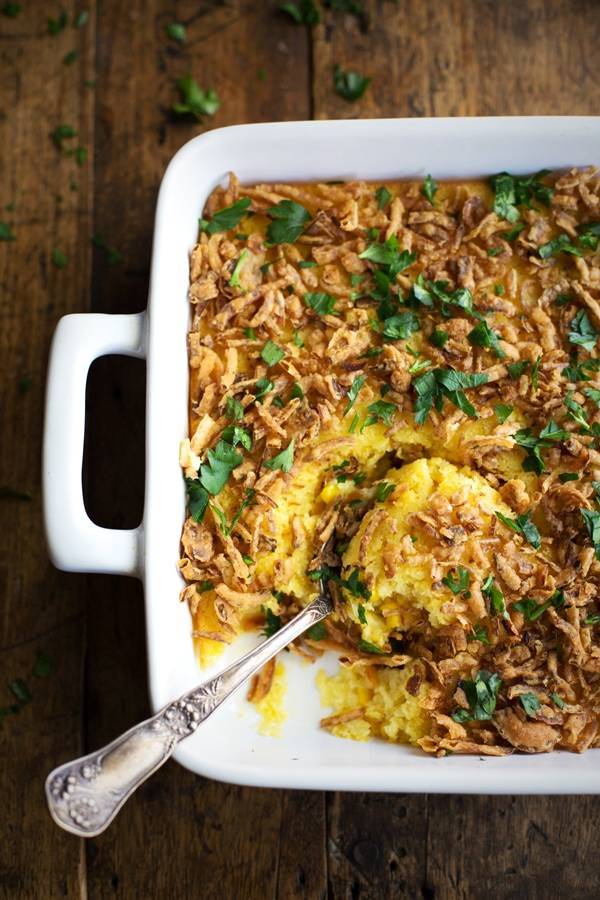 Corn pudding with crispy onions and herbs in a baking dish with a spoon.
