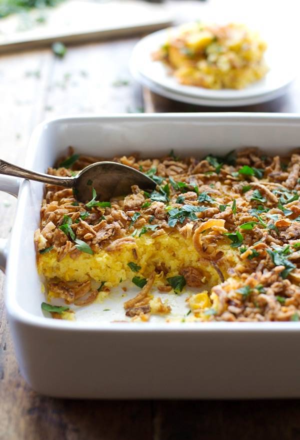 Creamy corn pudding with crispy onions and herbs in a white baking dish with a spoon.