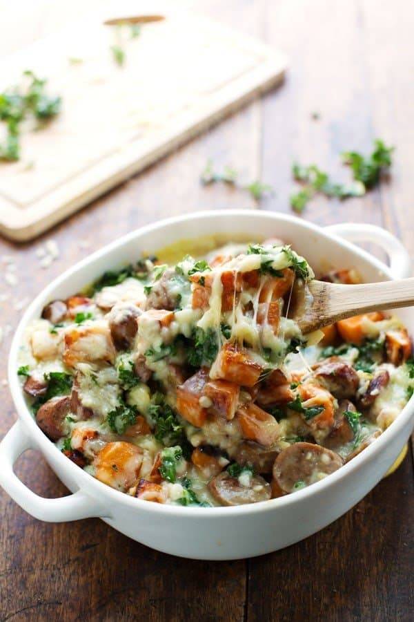 Sweet Potato, Kale, and Sausage Bake with White Cheese Sauce in a white baking dish with a wooden spoon.