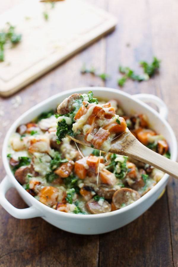 Sweet Potato, Kale, and Sausage Bake with White Cheese Sauce in a white baking dish and on a wooden spoon.