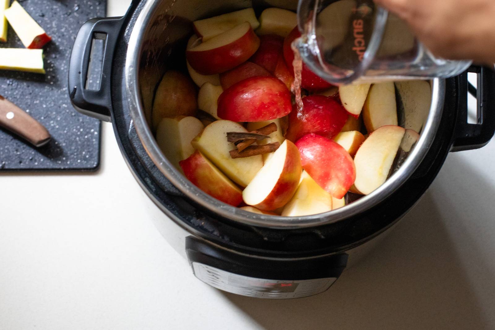Pouring water into an Instant Pot with apples and cinnamon sticks.
