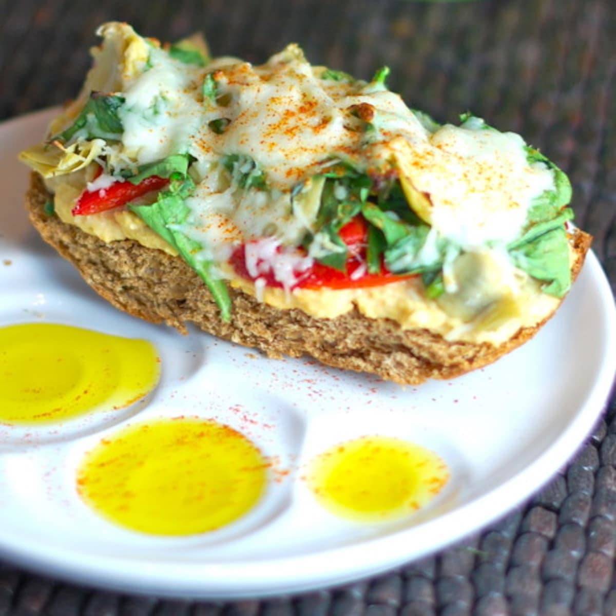 Open-faced sandwich with artichokes and red peppers over hummus and bread, drizzled with olive oil.
