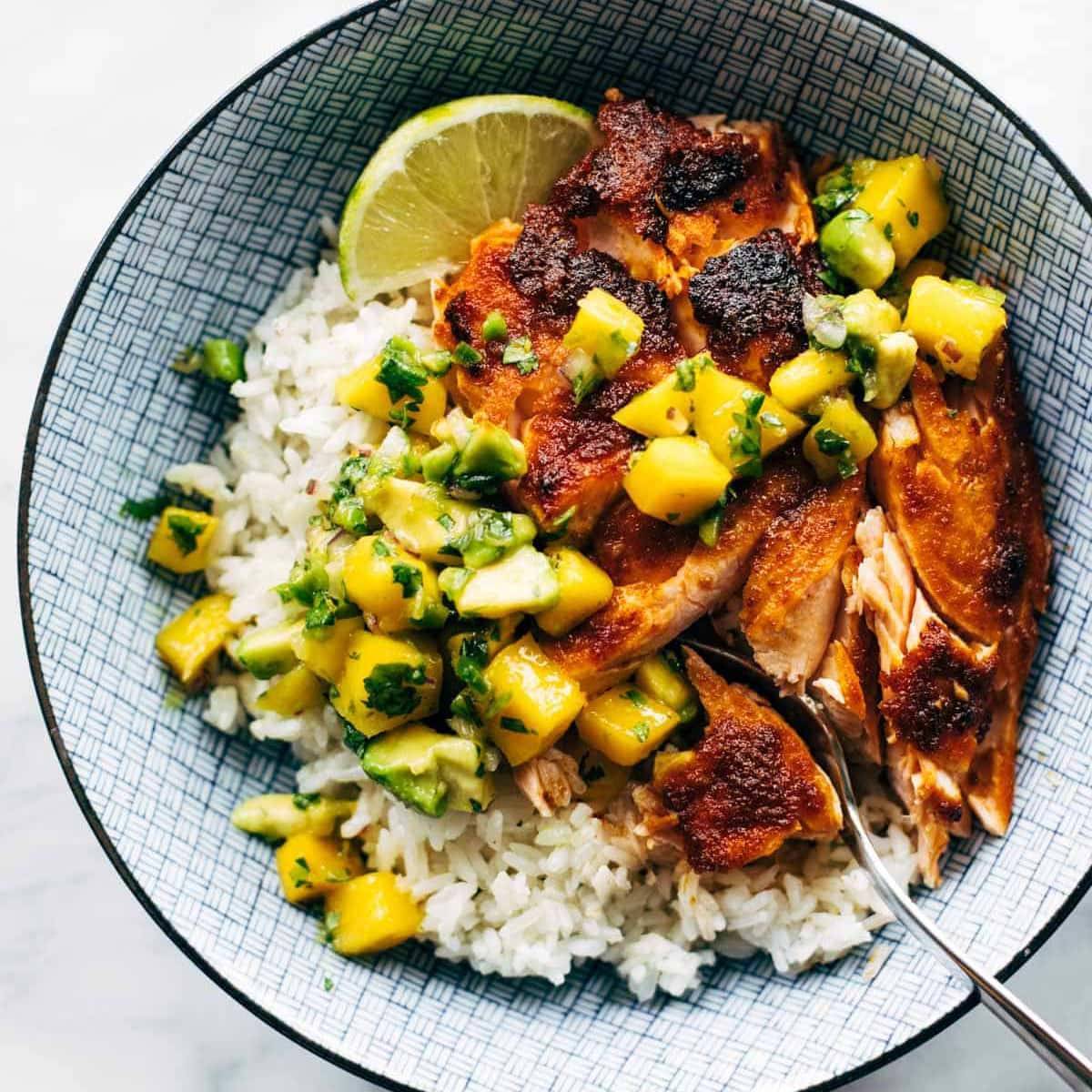 BBQ salmon and diced mango over white rice with a slice of lemon n a bowl with a spoon.