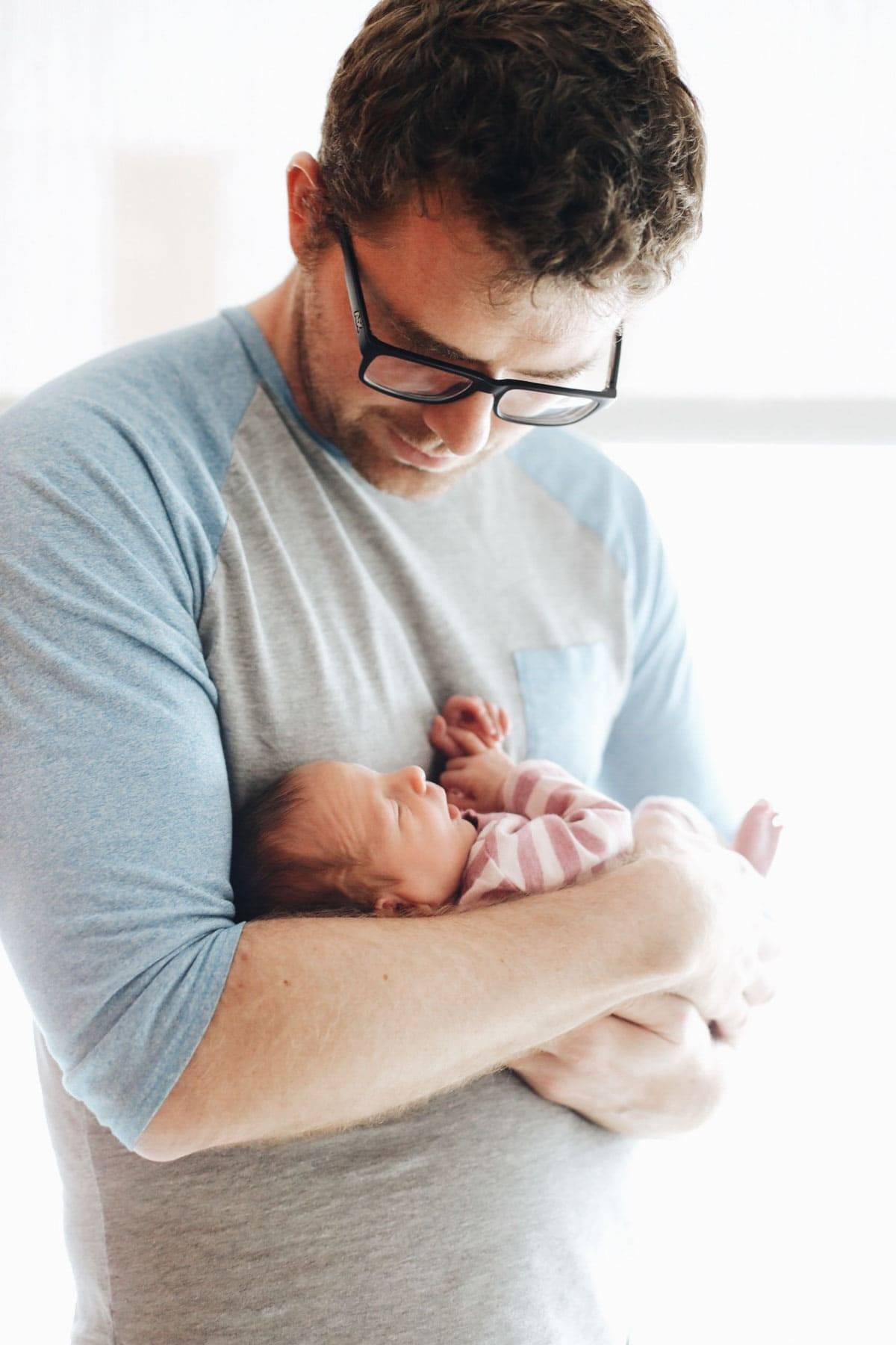 A man wearing glasses in a grayish blue shirt looking down on a tiny baby he is holding.