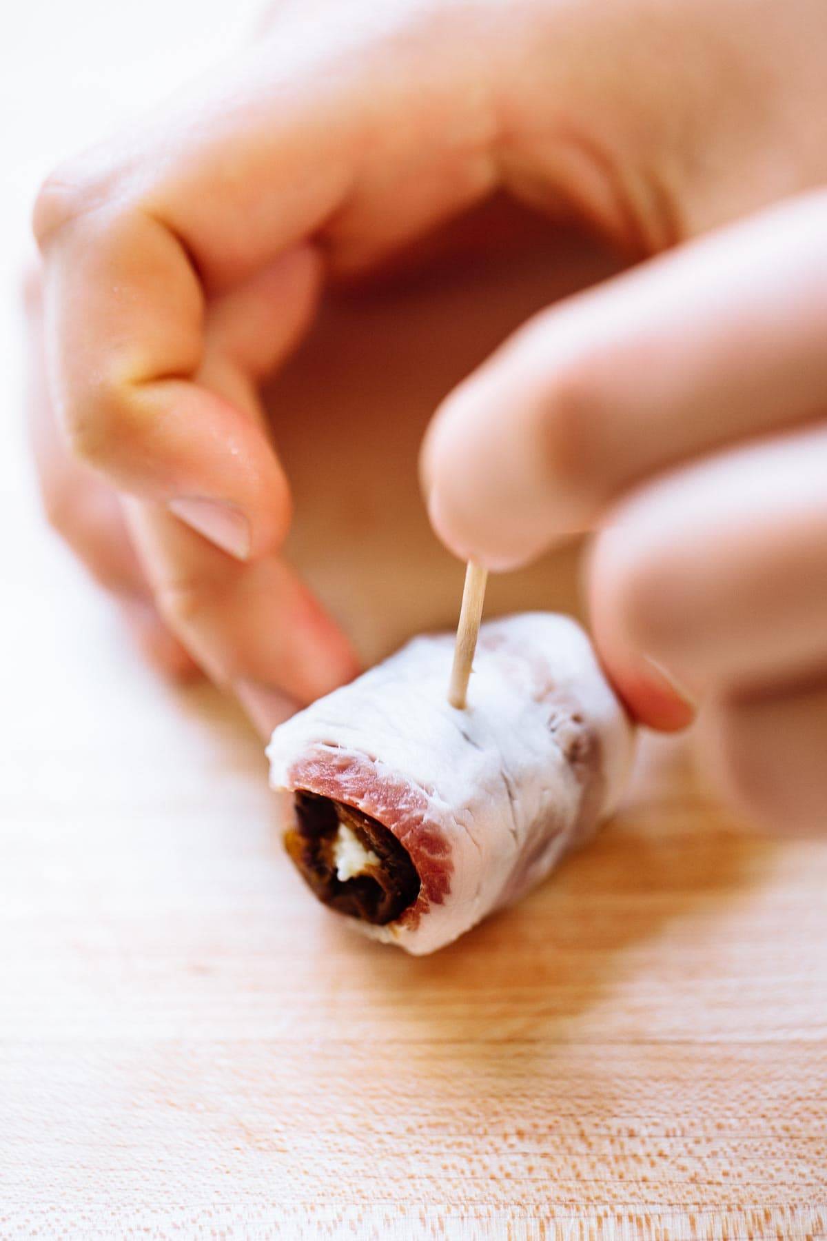 White hand sticking toothpick into date wrapped with bacon and stuffed with goat cheese. 