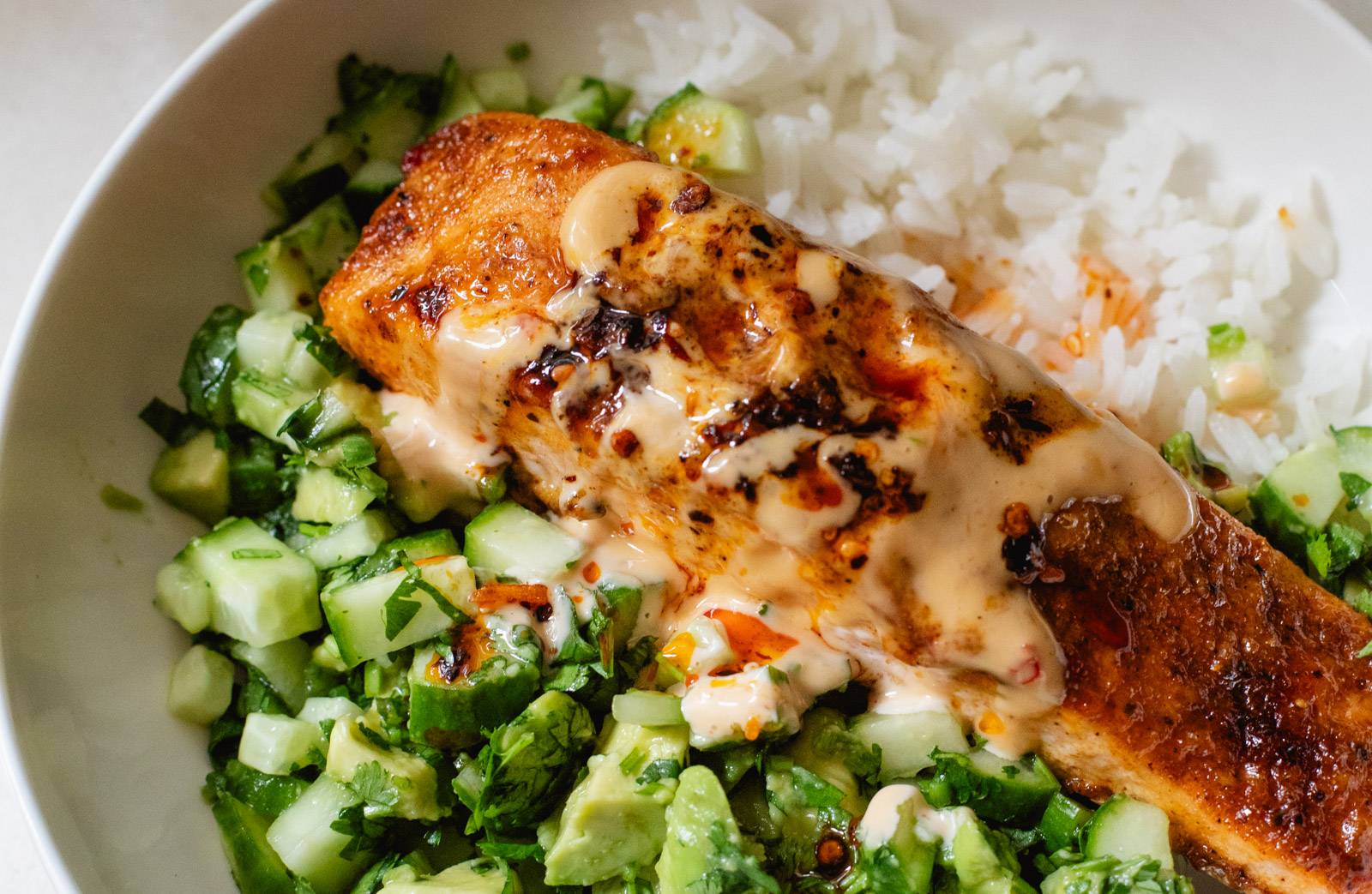 Bang bang salmon in a bowl with white rice and avocado cucumber salsa.