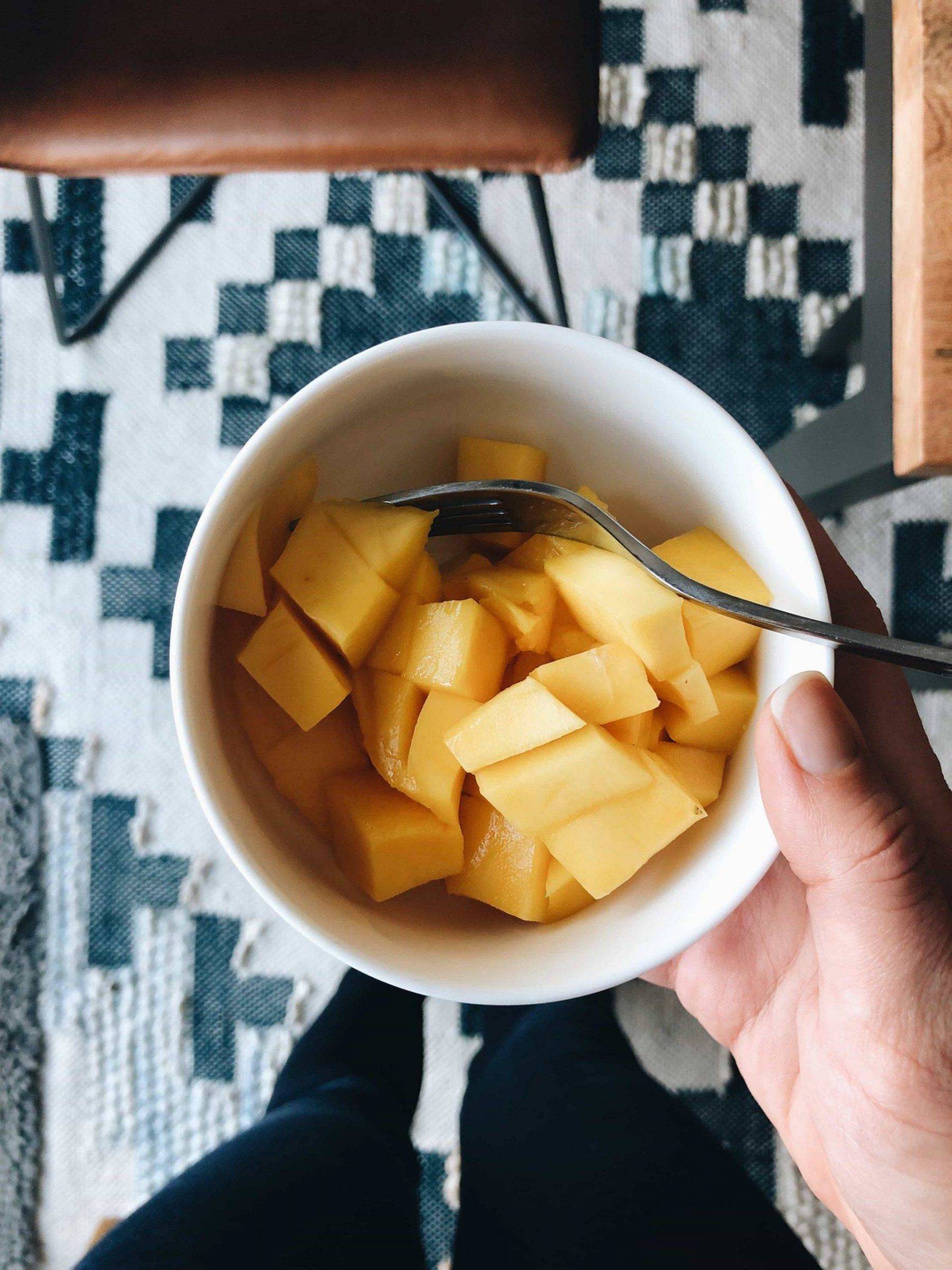 A bowl filled of fresh and juicy yellow colored mango pieces with a spoon in it and someone is holding the bowl.