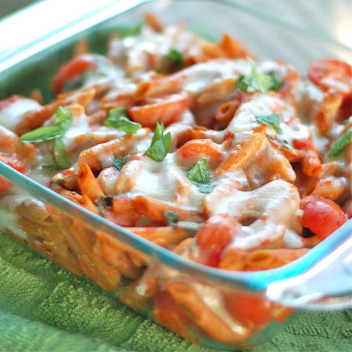 Vegetarian caprese penne pasta with sweet cherry tomatoes, mozzarella, and fresh basil in a clear baking dish.
