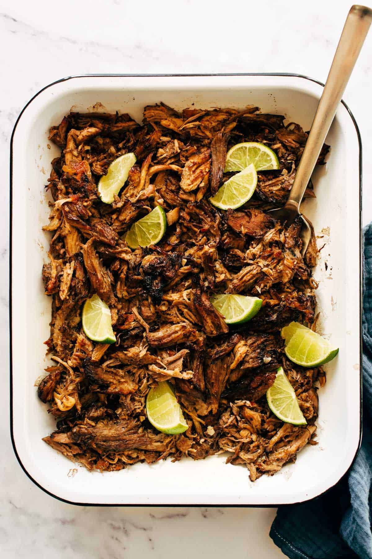 Carnitas in a pan with limes and a serving fork. 