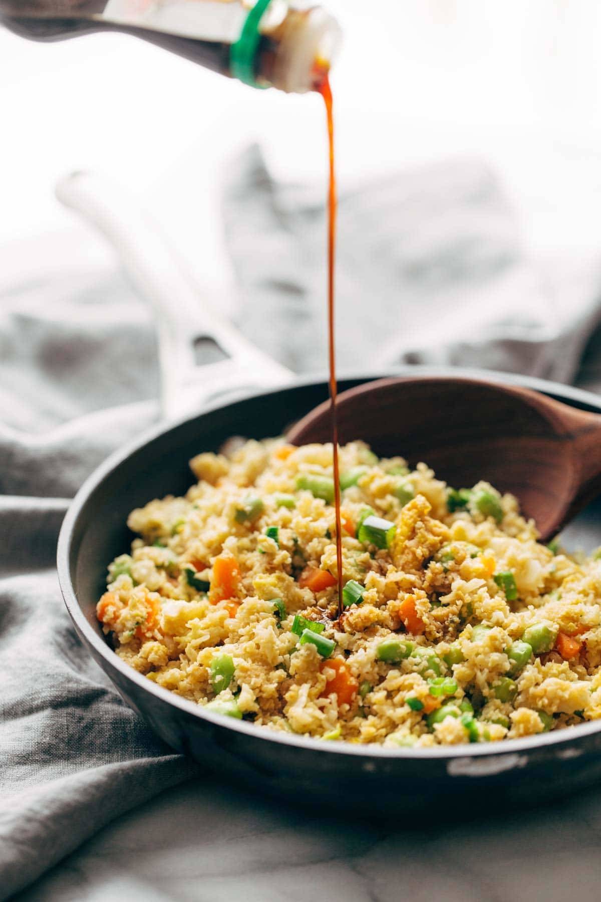 cauliflower fried rice in a bowl with a wooden spoon and a drizzle of soy sauce