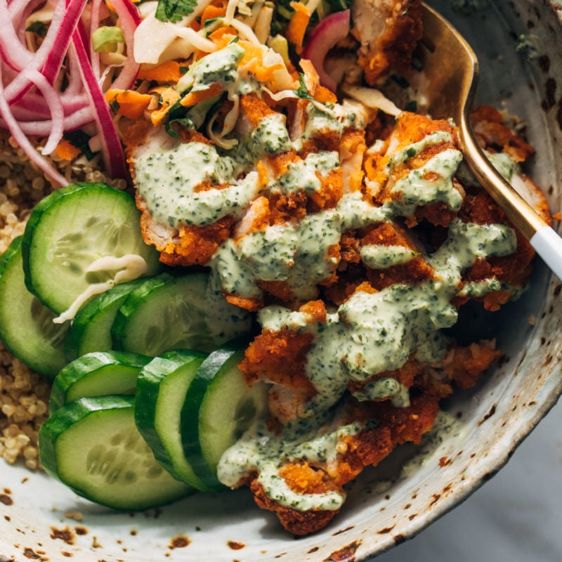 Crispy chicken, cucumbers, quinoa, and pickled red onions in a bowl with mint sauce and a fork