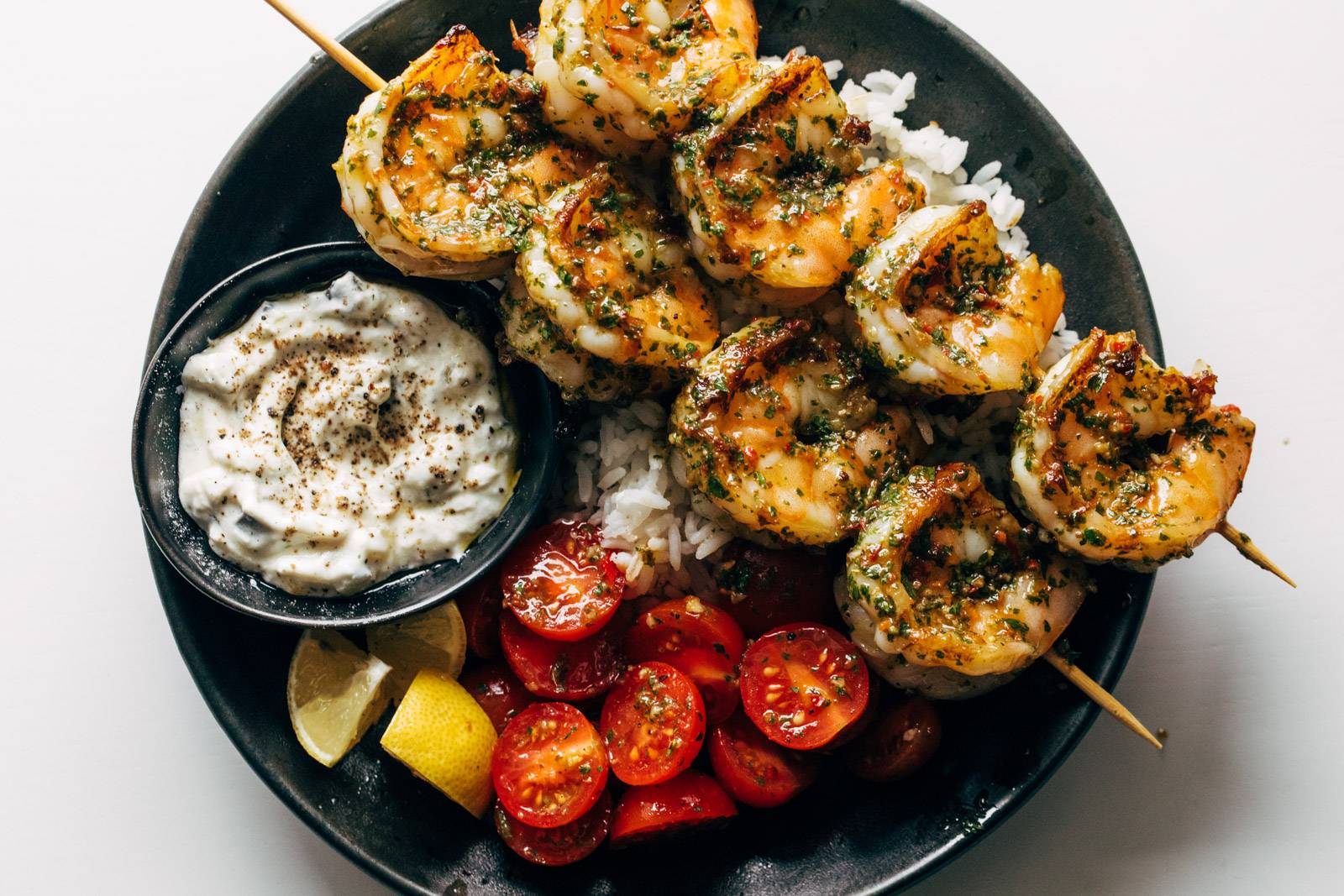 A plate filled with shrimp on skewers, rice, tomato salad, tzatziki, and lemon wedges.