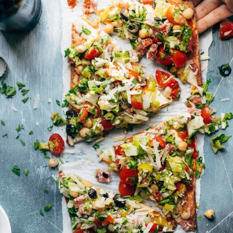 Olives, chickpeas, tomatoes, greens, herbs, and cheese on a thin crust flatbread pizza on a white plate.