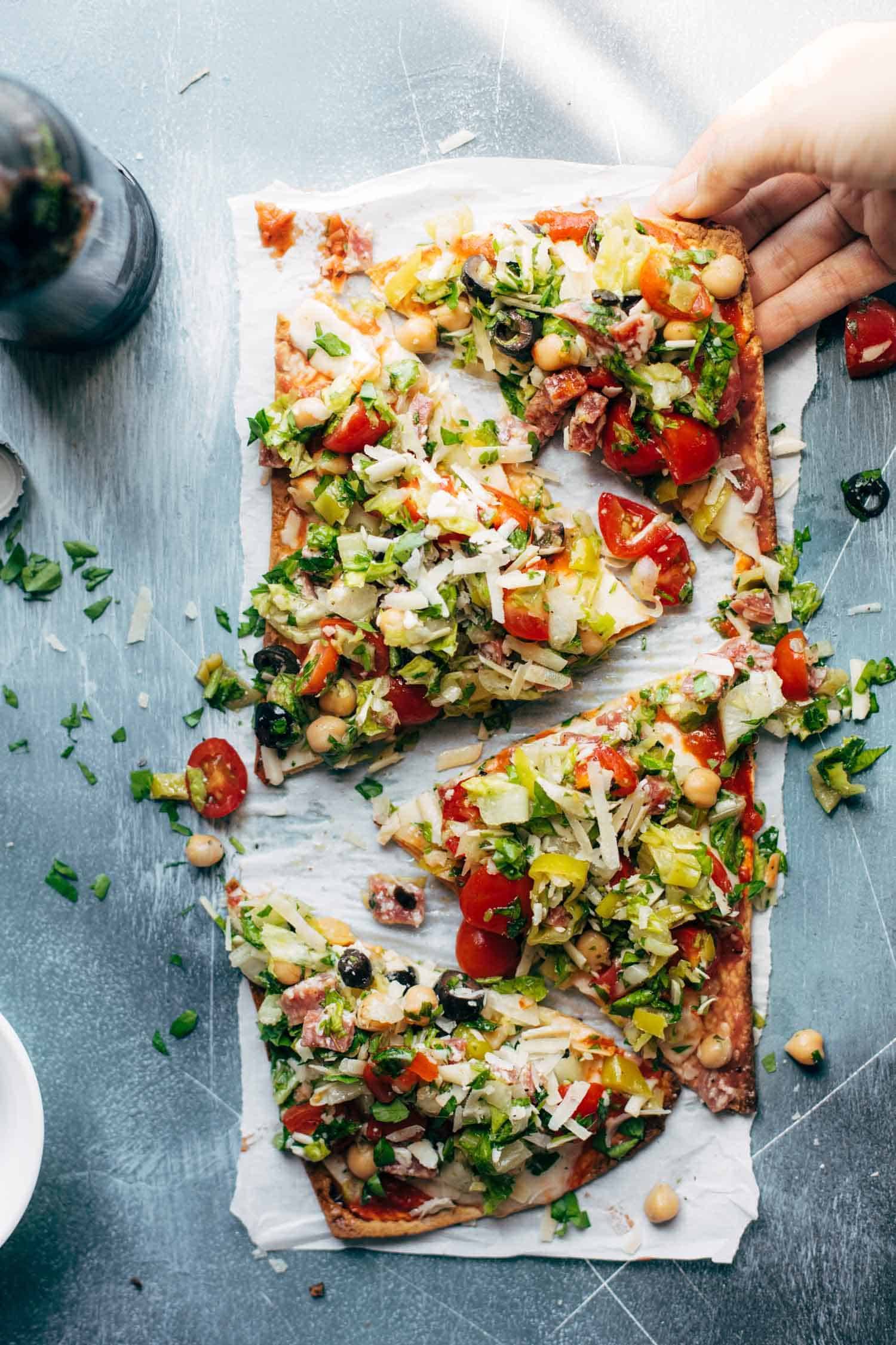 Olives, chickpeas, tomatoes, greens, herbs, and cheese on a thin crust flatbread pizza on a white plate.