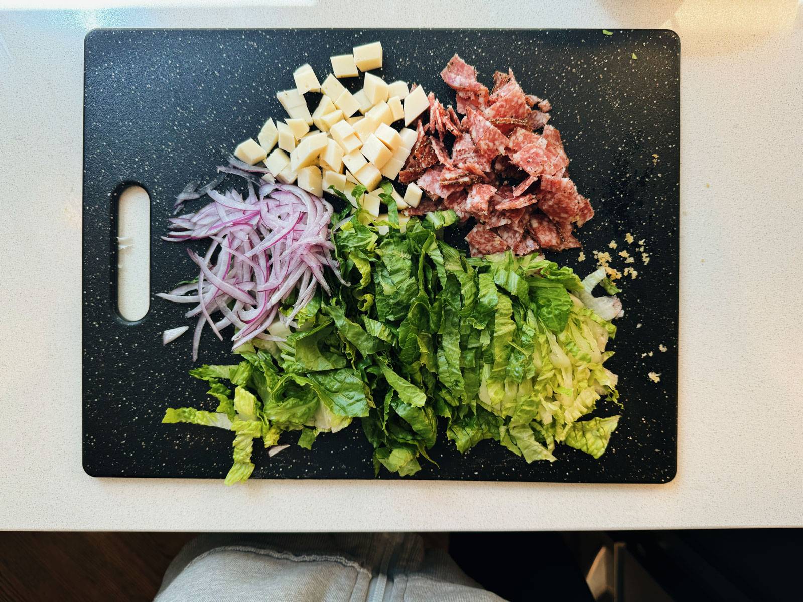 Chopped salad ingredients on a cutting board - romaine, red onion, cheese, and salami.