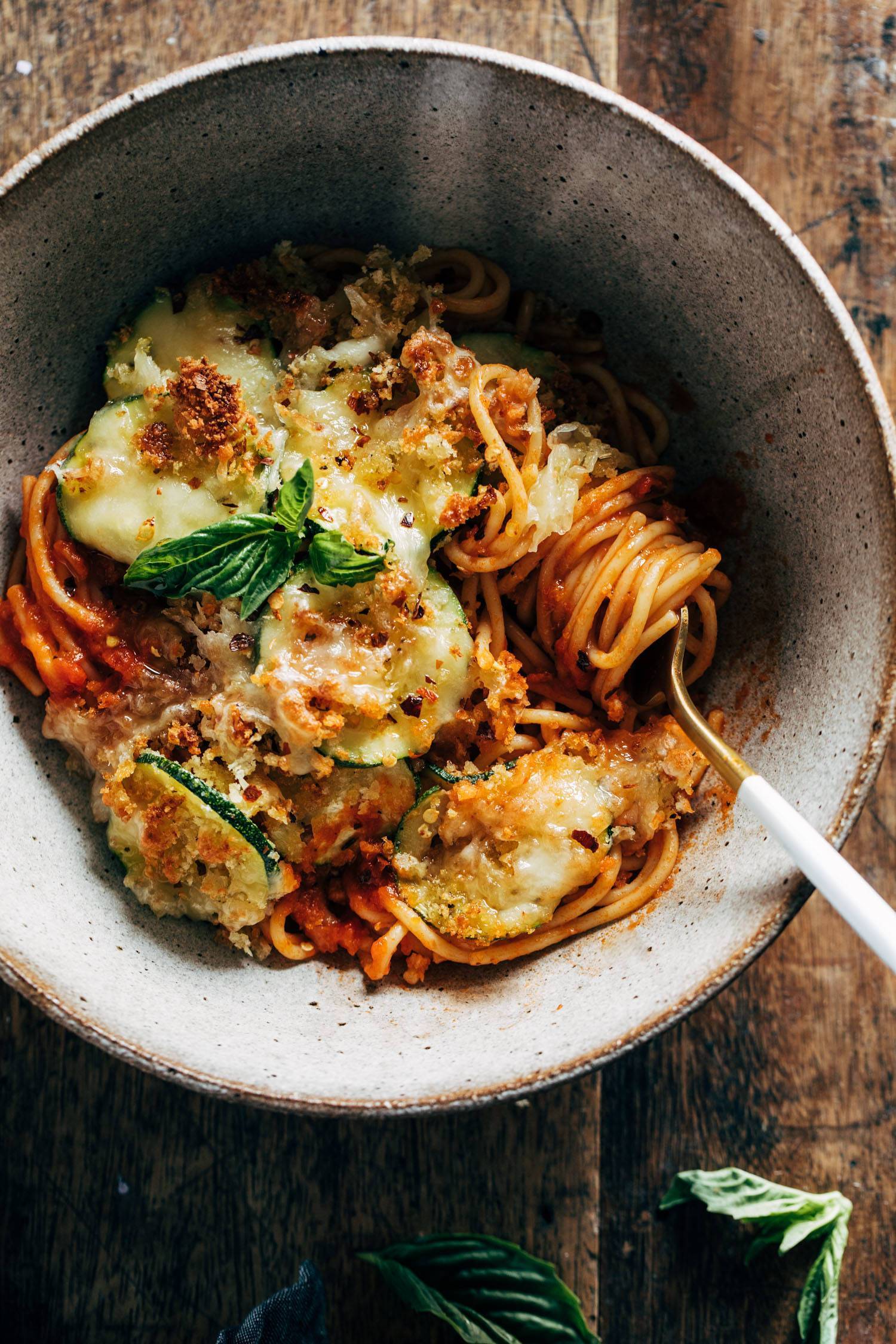 Spaghetti with crispy zucchini covered in melted cheese and fresh herbs in a bowl with a fork. 