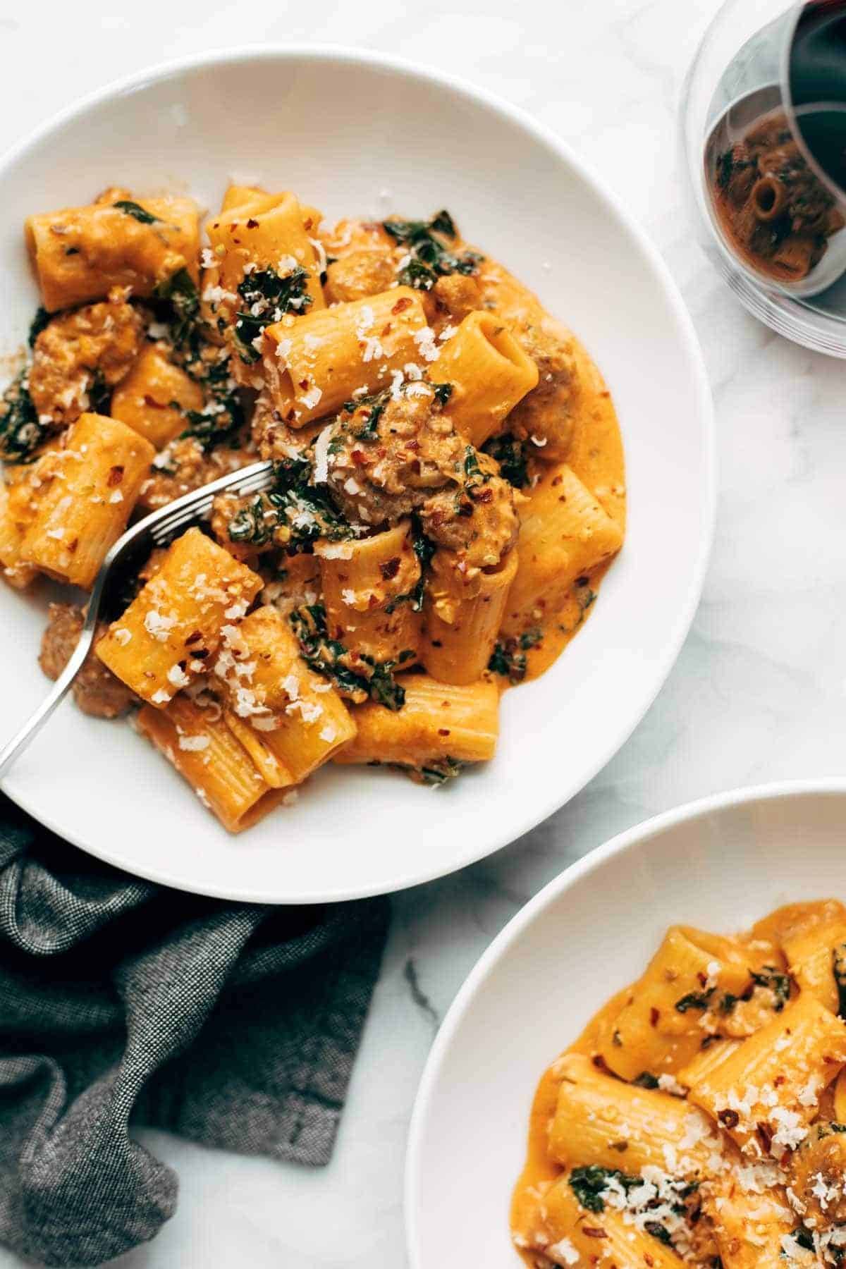 Rigatoni with sausage and kale in a bowl with a fork. There is a glass of red wine on the side. 