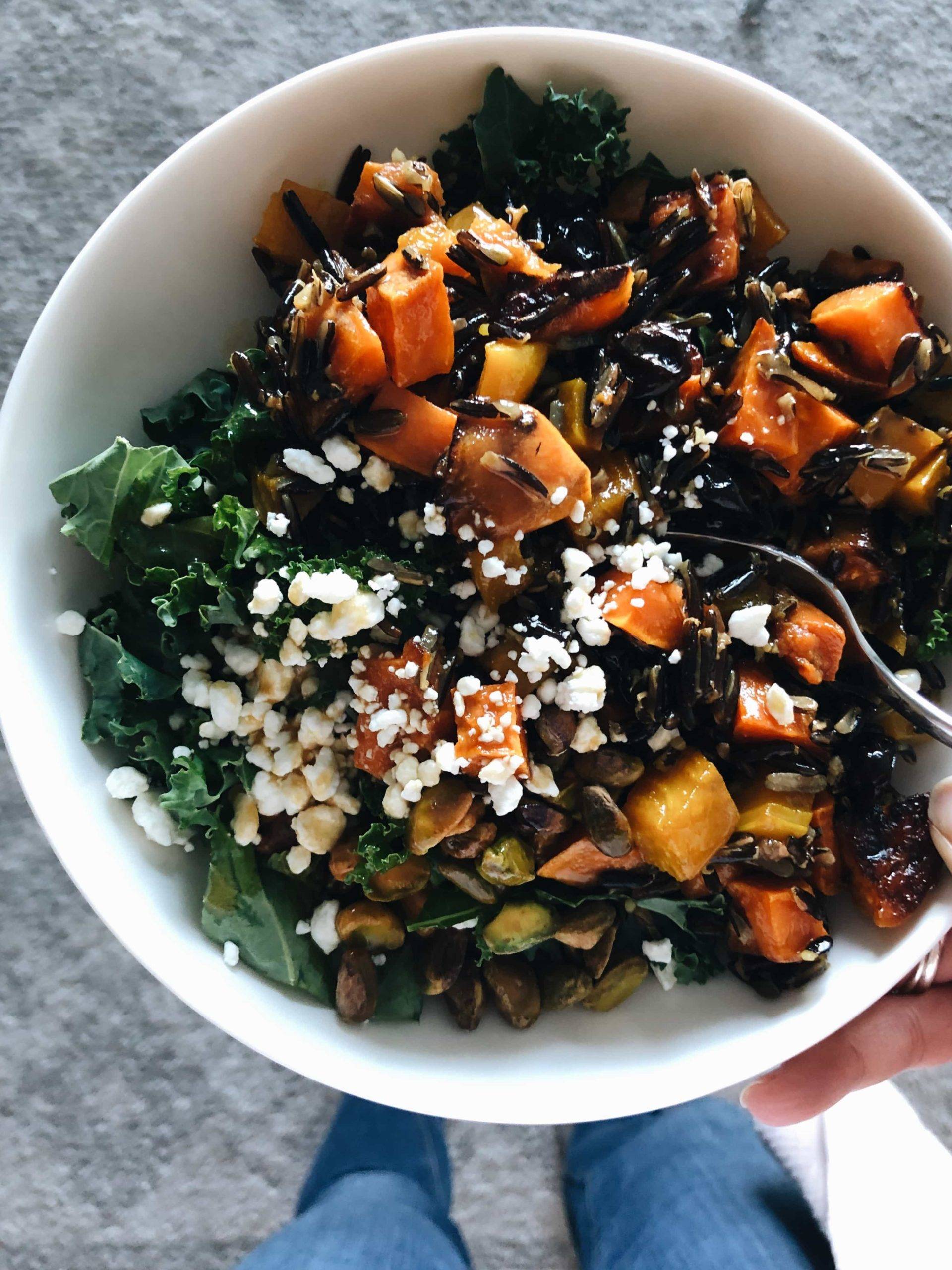 A large white bowl of squash, wild rice, pumpkin seeds, and feta cheese crumbles over wilted greens.