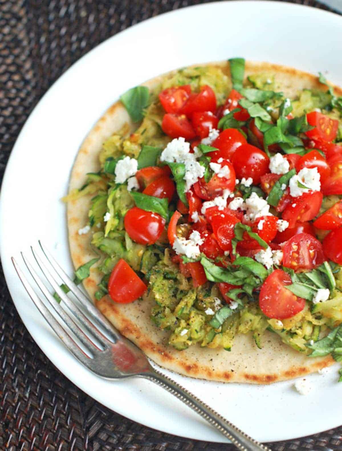 Flatbread pizza with zucchini, cherry tomatoes, and basil on a plate with a fork.