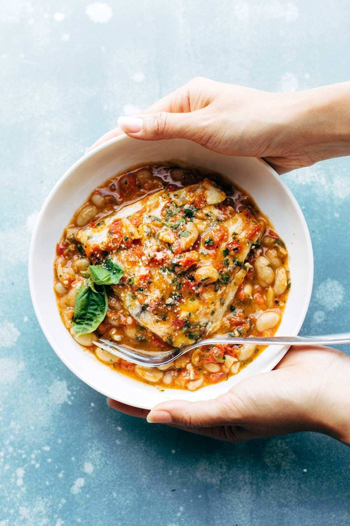 Hands Holding Garlic Basil Barramundi Skillet with Tomato Butter Sauce in a bowl with a fork.
