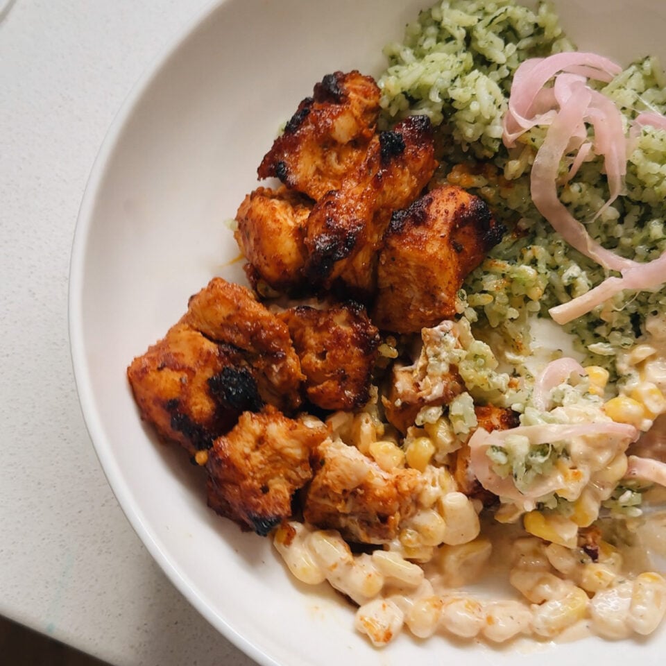 Green rice, chicken skewers, and creamy corn in a bowl.