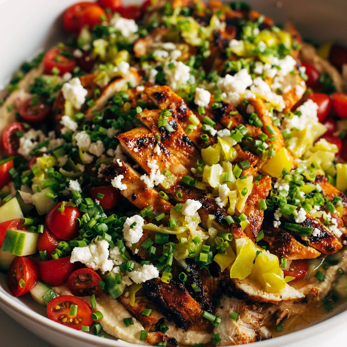 Grilled chicken in a bowl with hummus, herbs, and cheese.