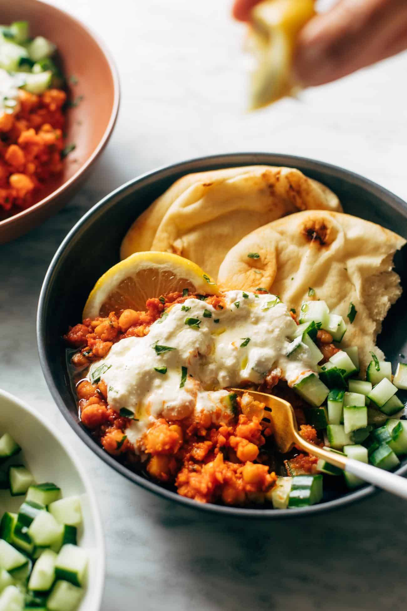 Harissa chickpeas with whipped feta in a bowl with cucumbers, a fork, lemon, and naan. 