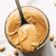 Peanut butter in a jar with a spoon and peanuts scattered around.