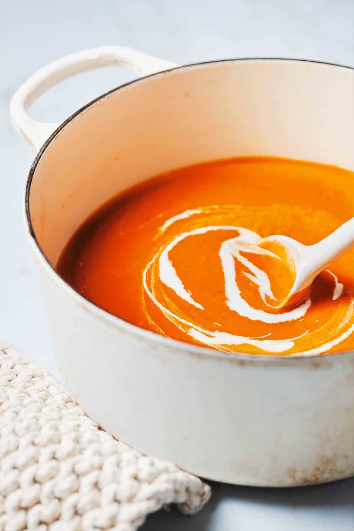 Tomato soup with cream being stirred in a white pot with a white spoon.