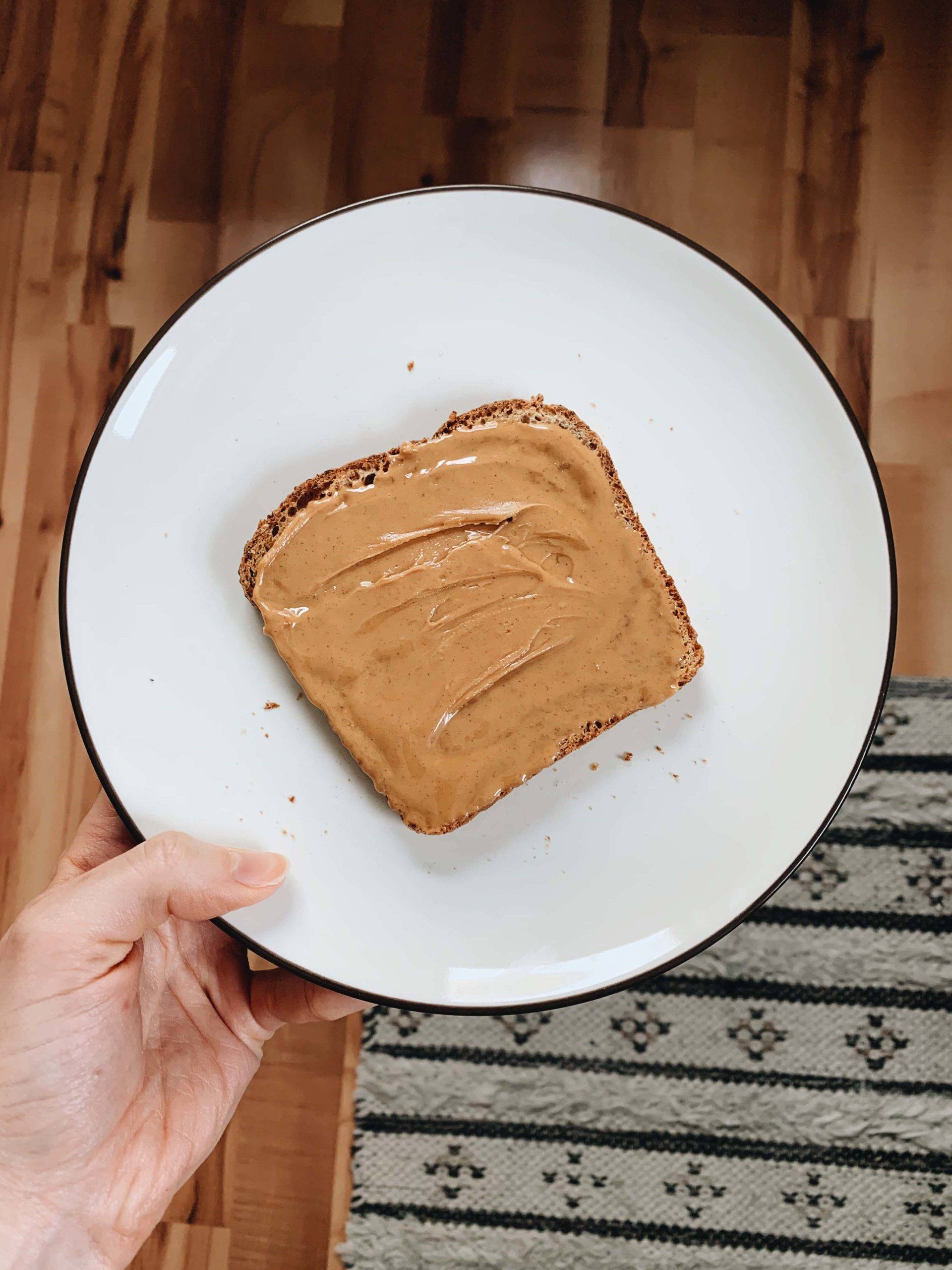 A hand holding a white plate with a piece of bread covered with peanut butter on the plate.