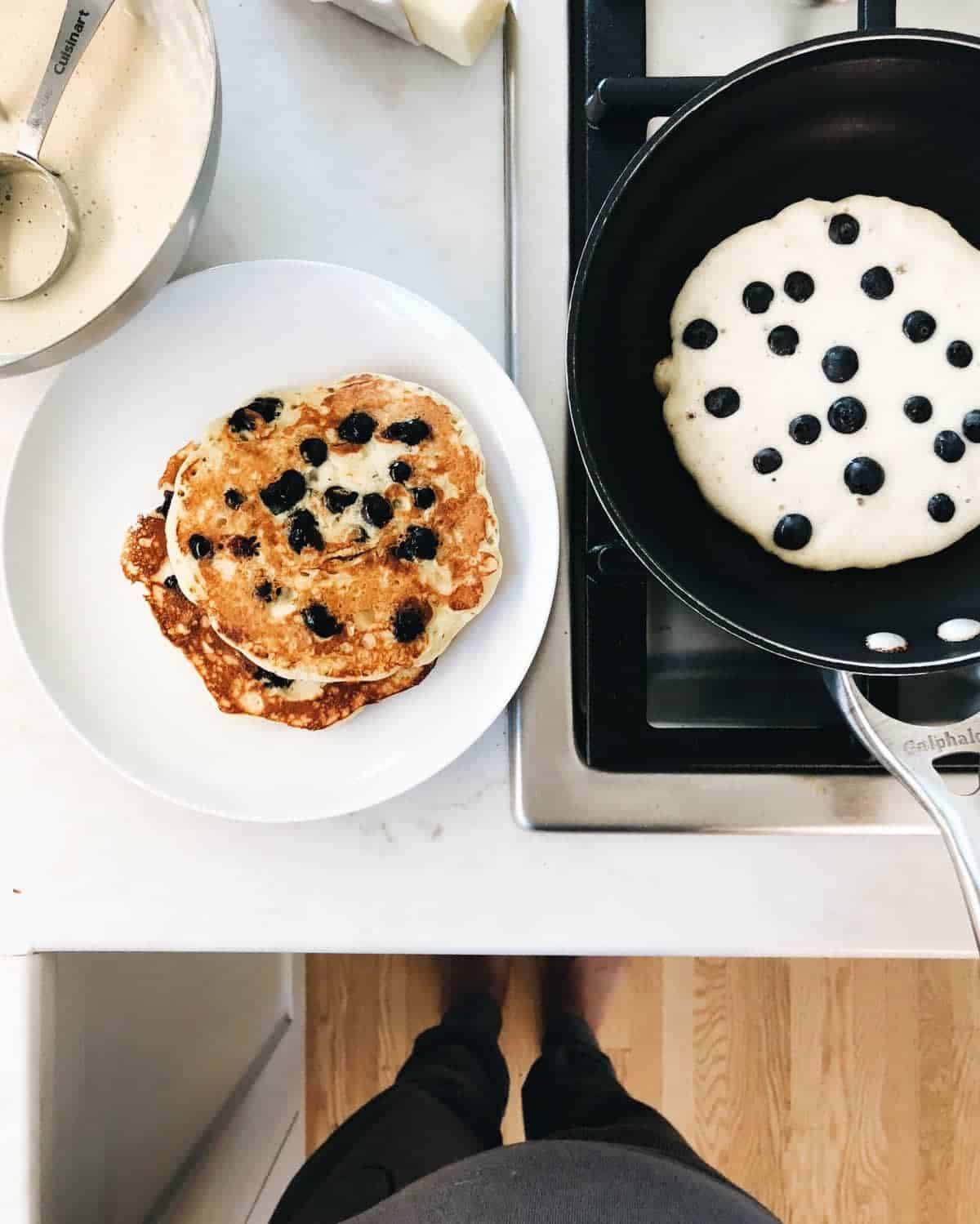 A large white plate of blueberry pancakes with butter melting over them as some cook in a skillet next to it.