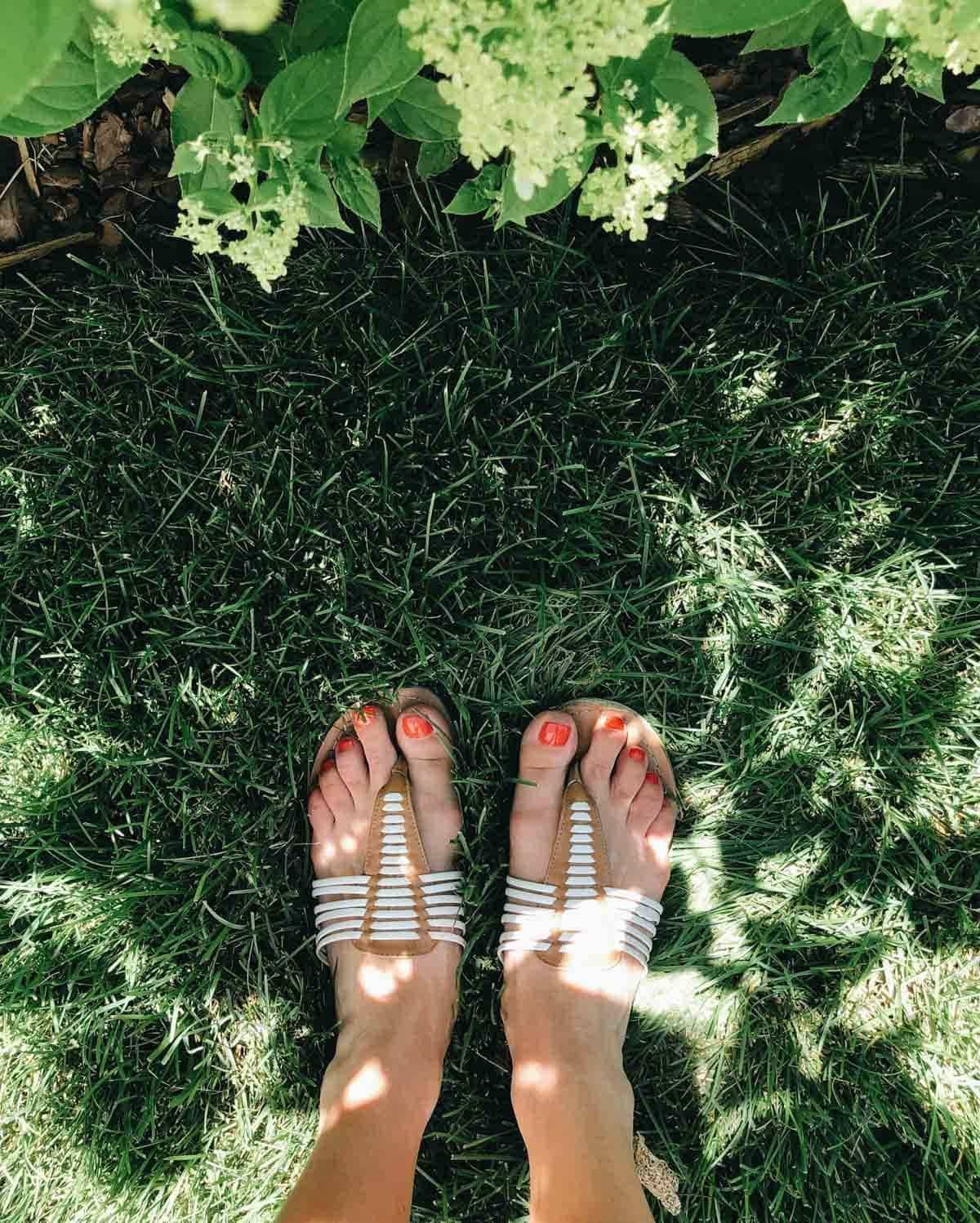 Feet of a woman having red nail polish, wearing sandals can be seen on the green grass.
