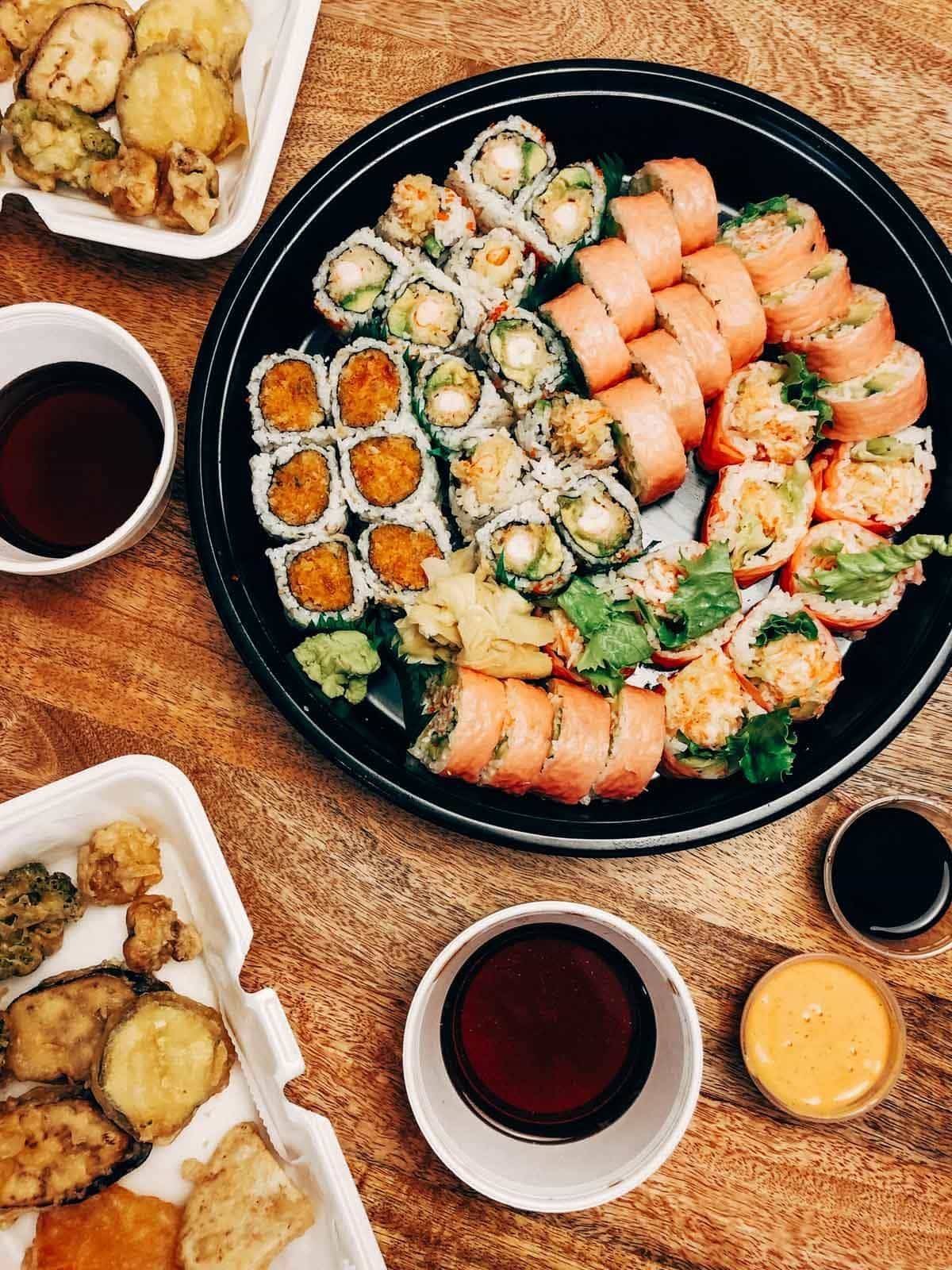 A wooden table is filled with large plate of sushi with sauces and drinks.