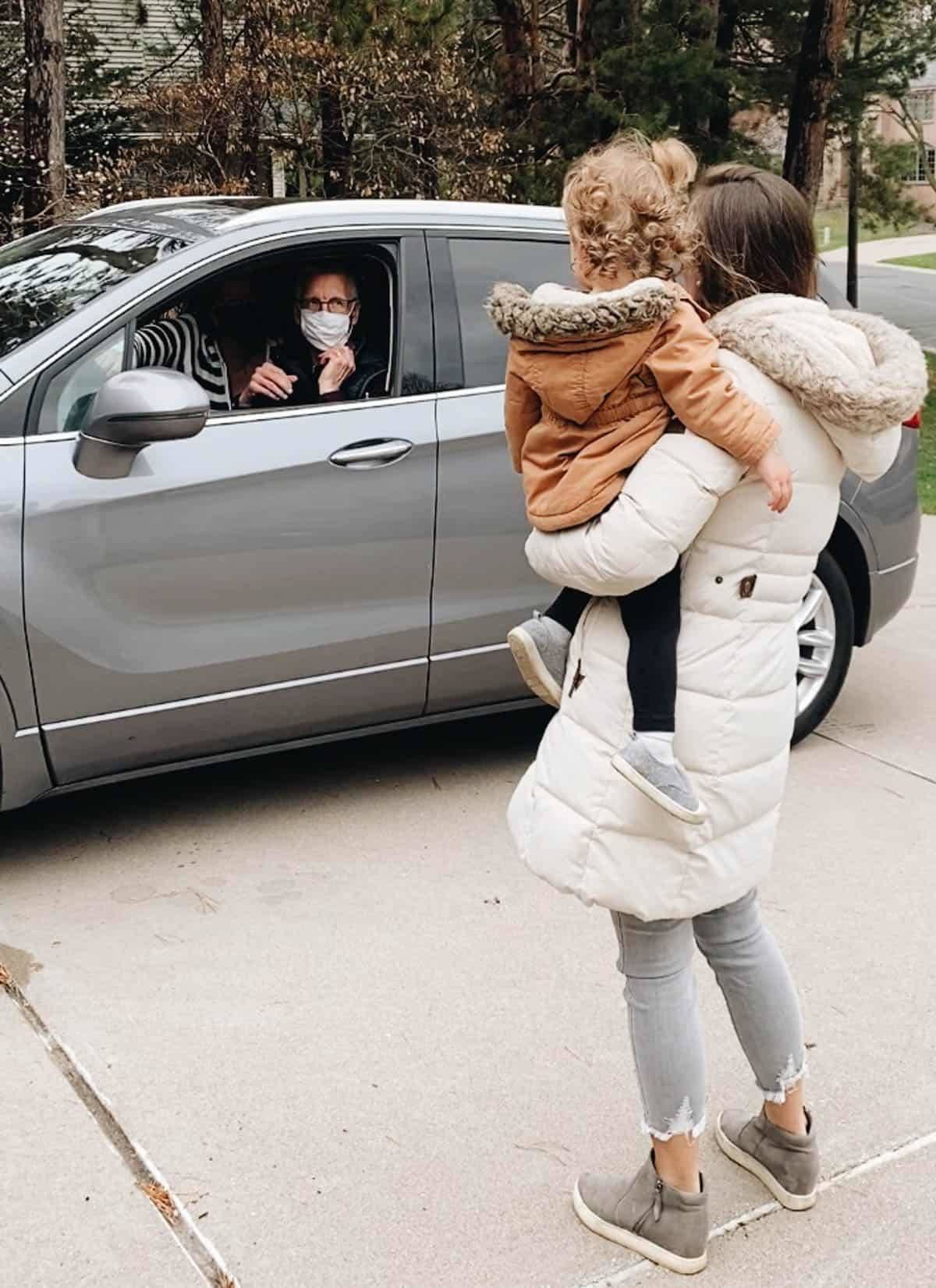 A woman holding a child on her hip as she talks to someone in a grey car.