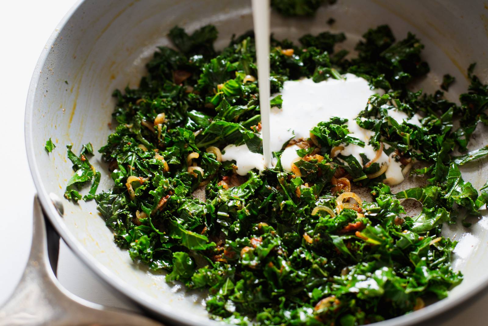 Pouring coconut milk into a pan of sautéed kale.