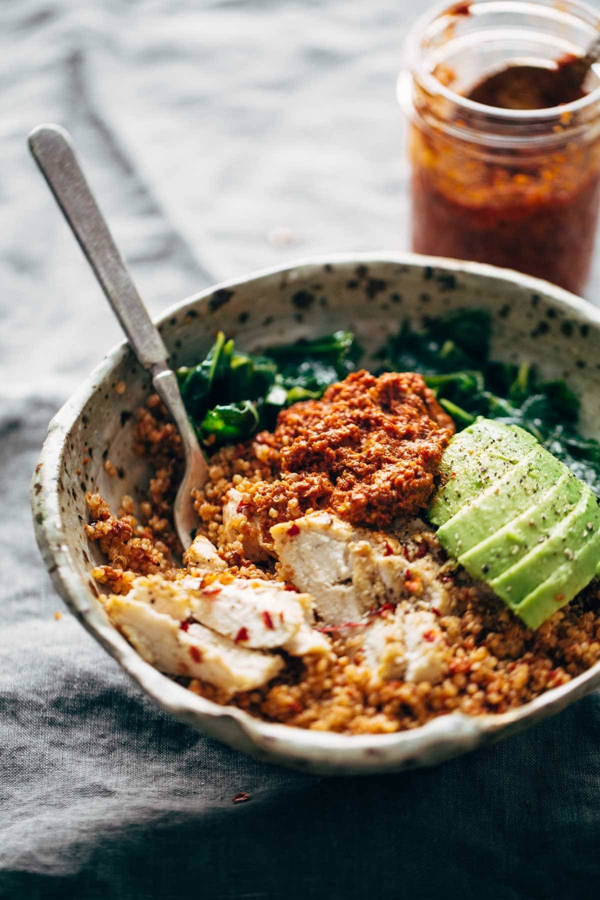 bowl with kale, chicken, quinoa and sun dried tomatoes with a fork and jar of sauce