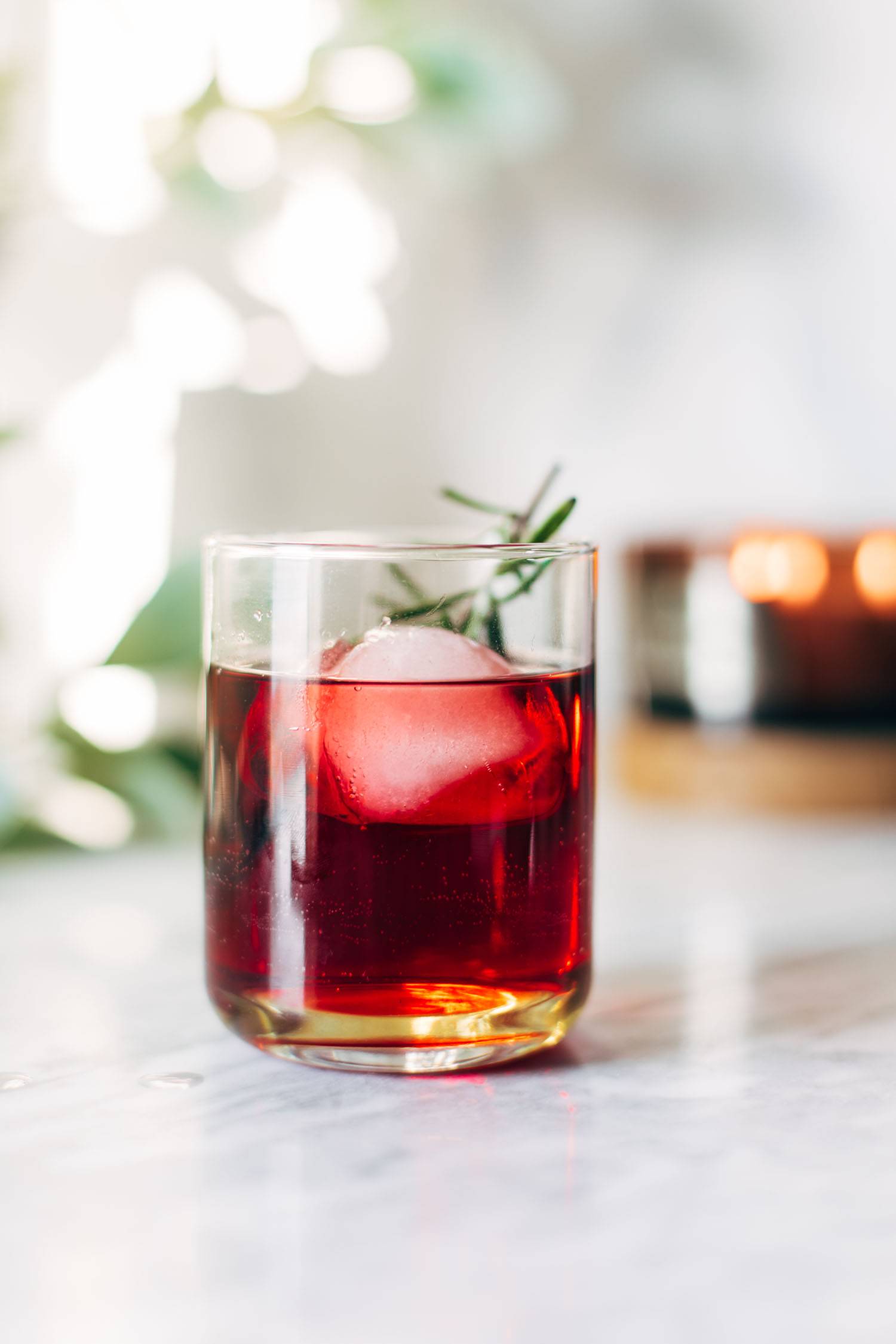 A clear cocktail glass with red liquid, a large ice cube, and a sprig of rosemary.