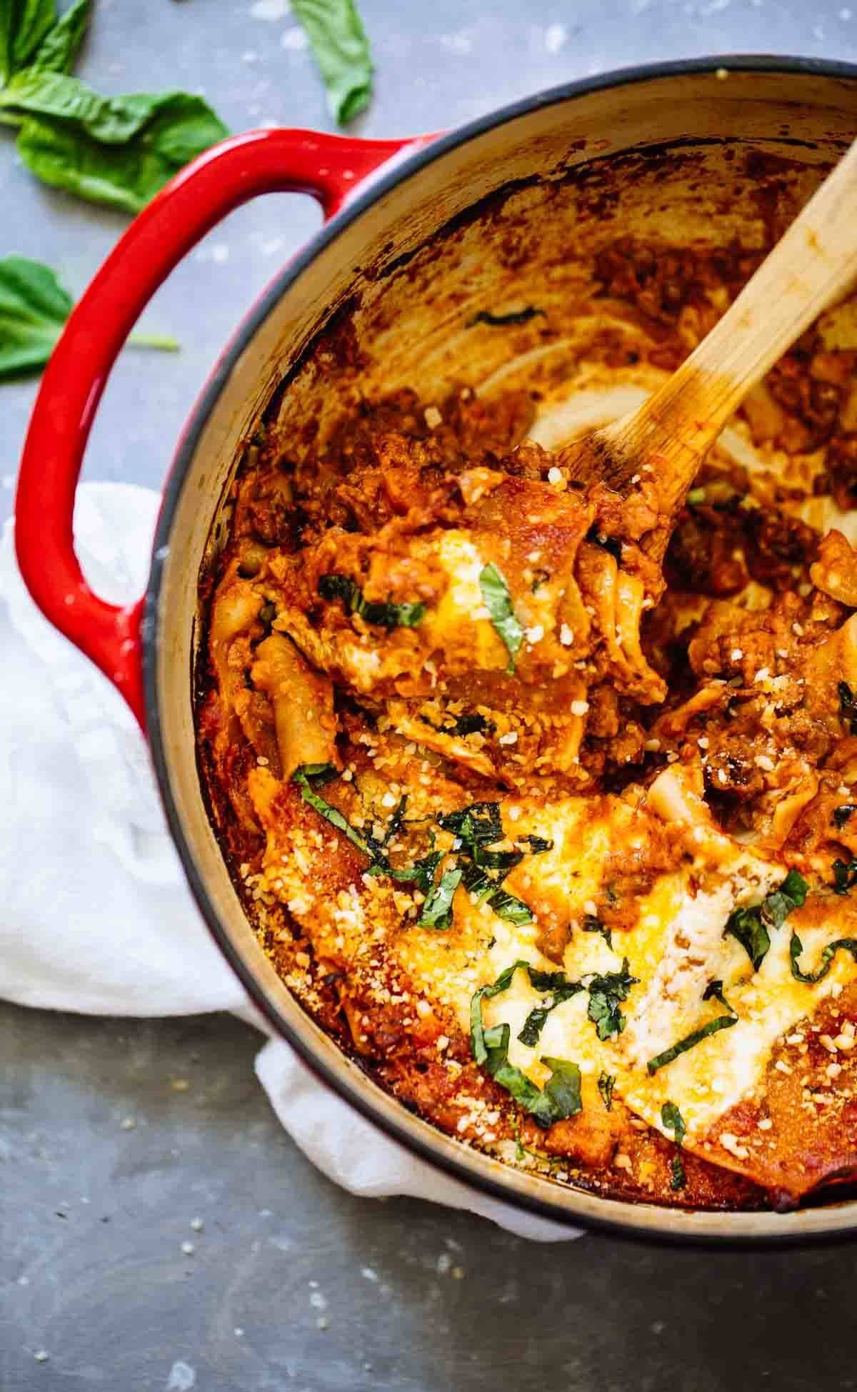 Lasagna in a red dutch oven with a wooden spoon.