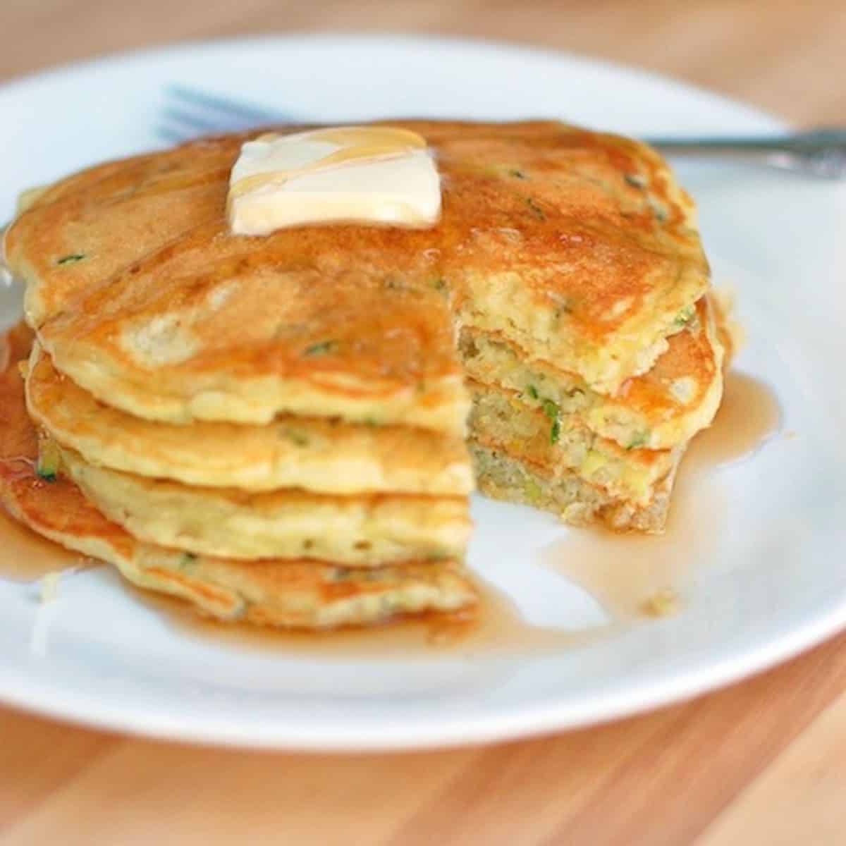 Stack of zucchini pancakes with butter and maple syrup on a white plate.