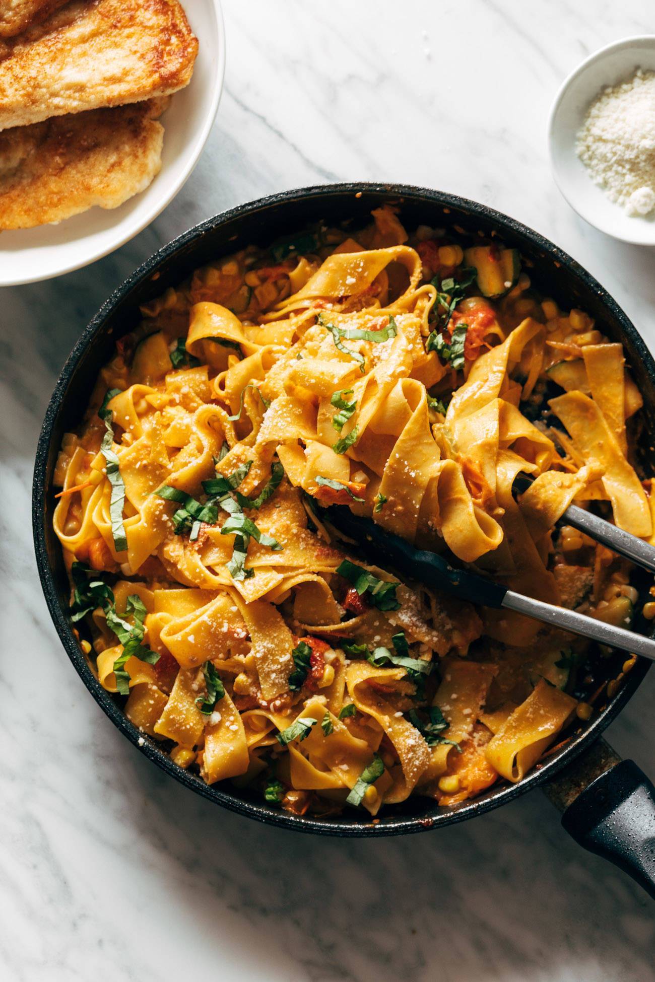 Burst tomato pappardelle in a pan topped with fresh basil and being scooped with tongs. 