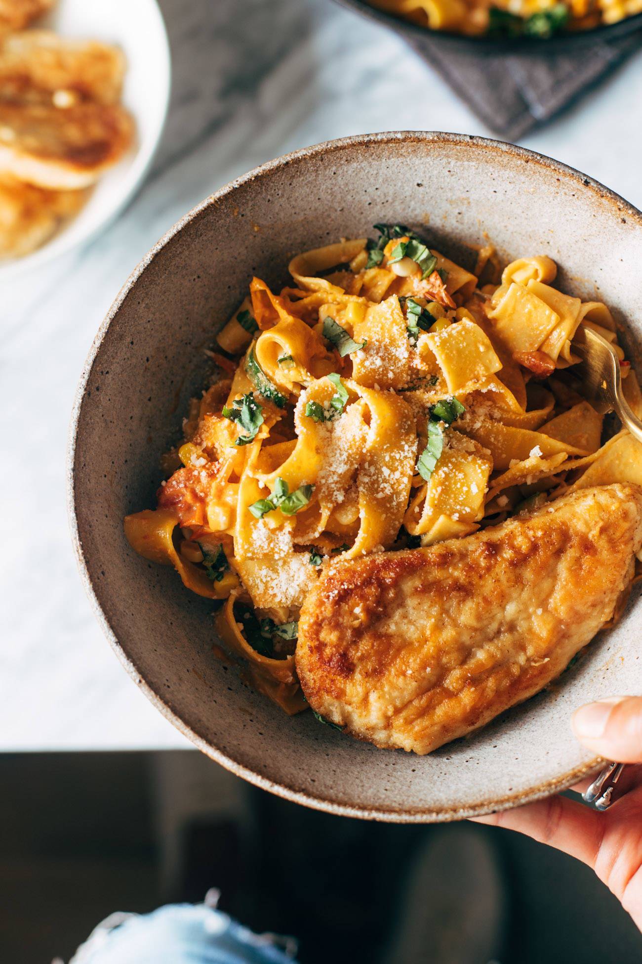 Burst tomato pappardelle in a bowl with a white hand holding the bowl. There's a pan-fried piece of chicken on top of the pasta. 