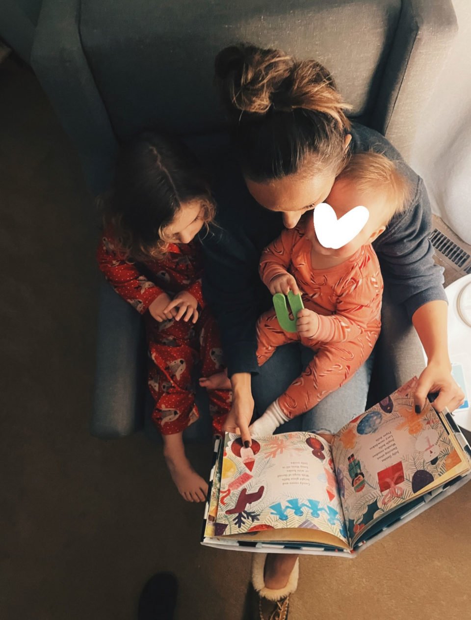 White woman reading a book to her two daughters  in a chair.