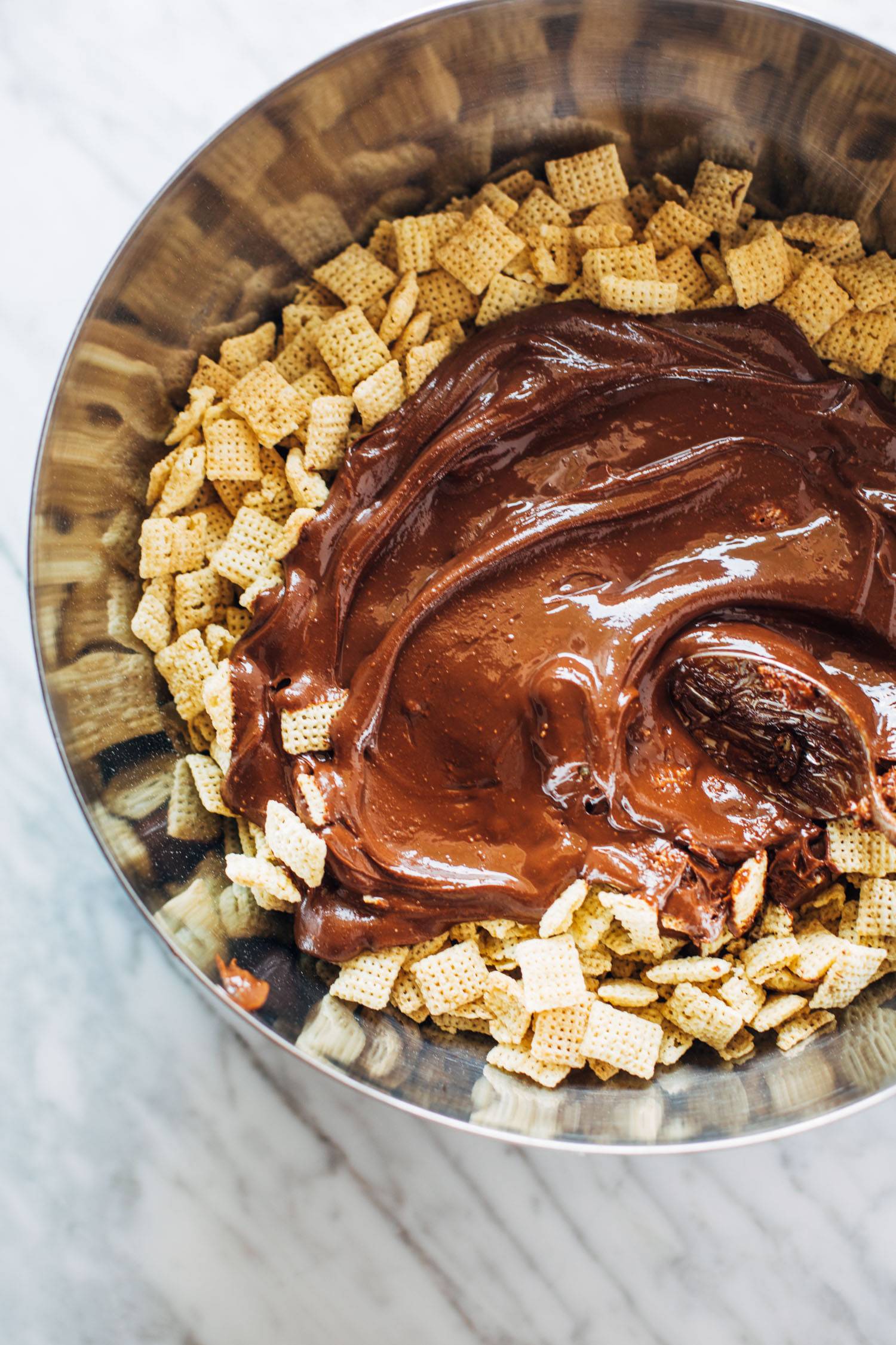 Melted chocolate being mixed into chex mix in a bowl.